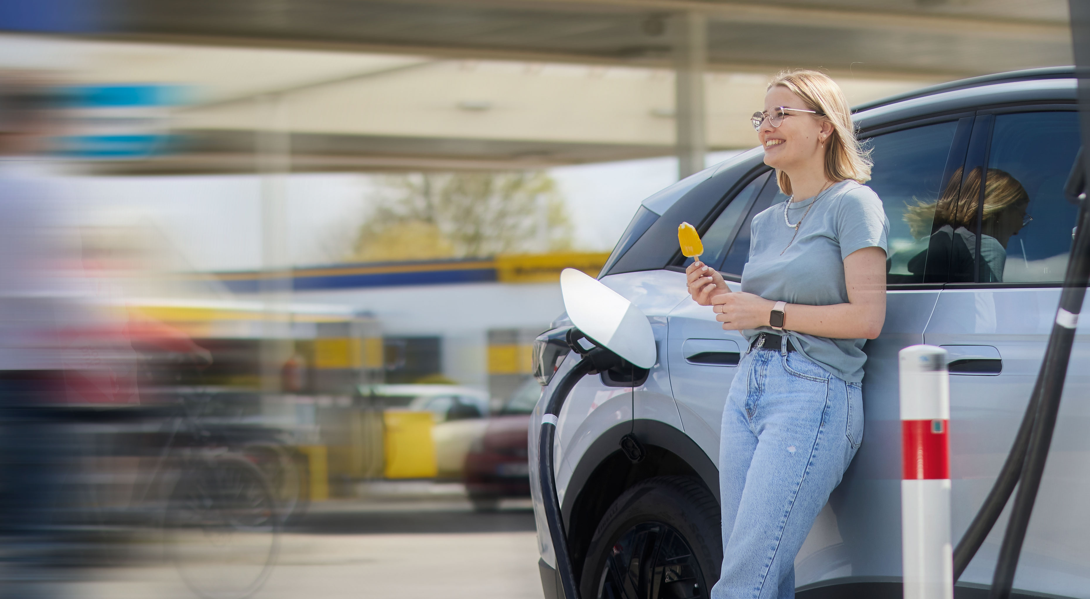 Frau isst Eis am Elektroauto während des Ladevorgangs an einer Westfalen Tankstelle